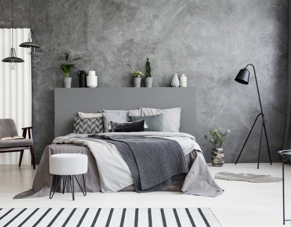 Grey armchair and stool near bed with headboard in bedroom interior with black lamp. Real photo