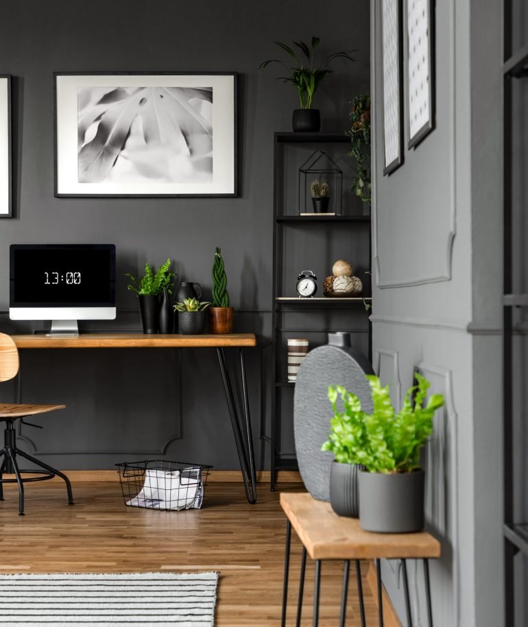 Plants on wooden table in grey home office interior with posters above desk with computer monitor