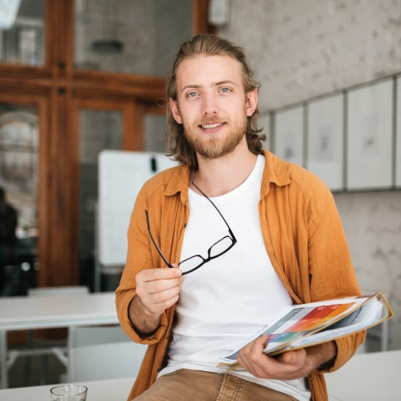 Young man sitting on table in office with glasses and document case in hands. Portrait of smiling boy with blond hair and beard joyfully looking in camera while holding documents in hand