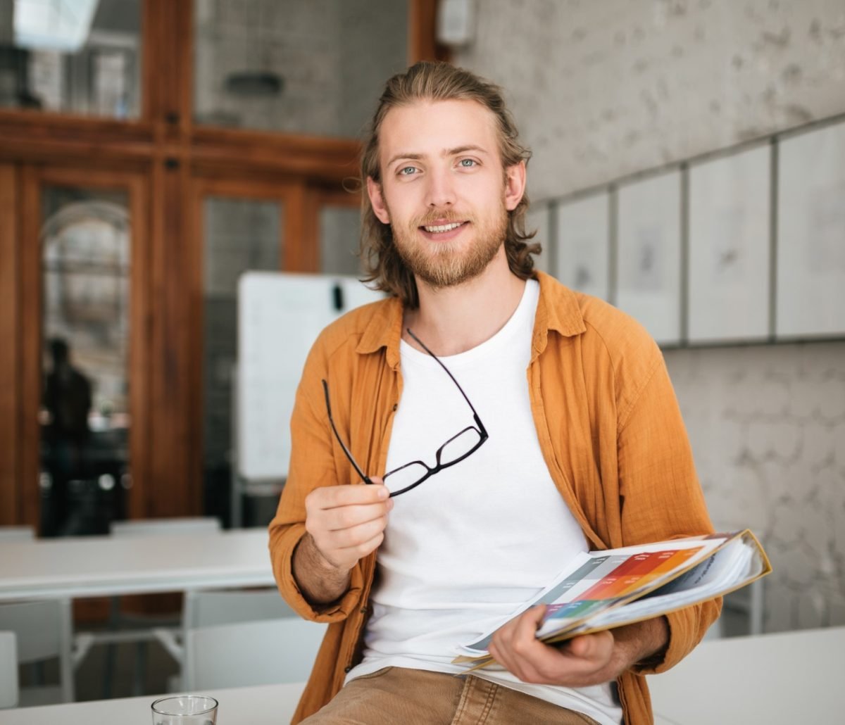Young man sitting on table in office with glasses and document case in hands. Portrait of smiling boy with blond hair and beard joyfully looking in camera while holding documents in hand