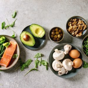 Ketogenic diet ingredients for cooking dinner. Raw salmon, avocado, broccoli, bean, olives, nuts mushrooms, eggs in ceramic bowls. Grey texture background. Flat lay, space