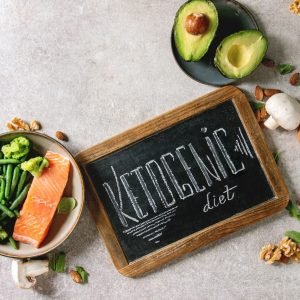 Ketogenic diet ingredients for cooking dinner. Raw salmon, avocado, broccoli, bean, olives, nuts, mushrooms, eggs in ceramic bowls with chalk board lettering. Grey texture background. Flat lay, space