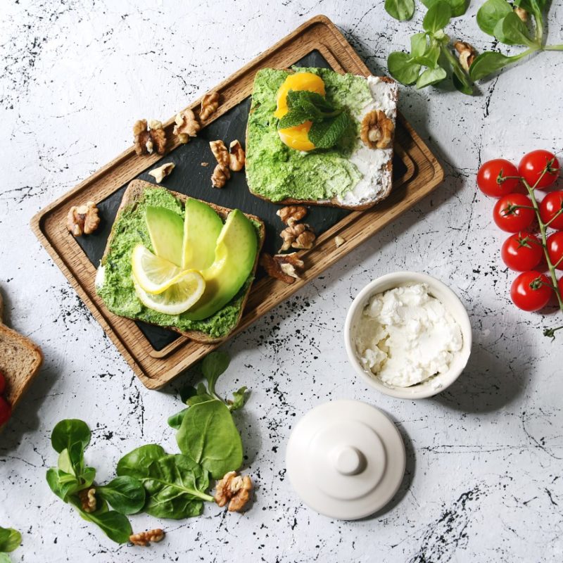 Vegetarian sandwiches with avocado, ricotta, egg yolk, spinach, cherry tomatoes on whole grain toast bread on wooden slate board with ingredients above over white marble background. Top view, space