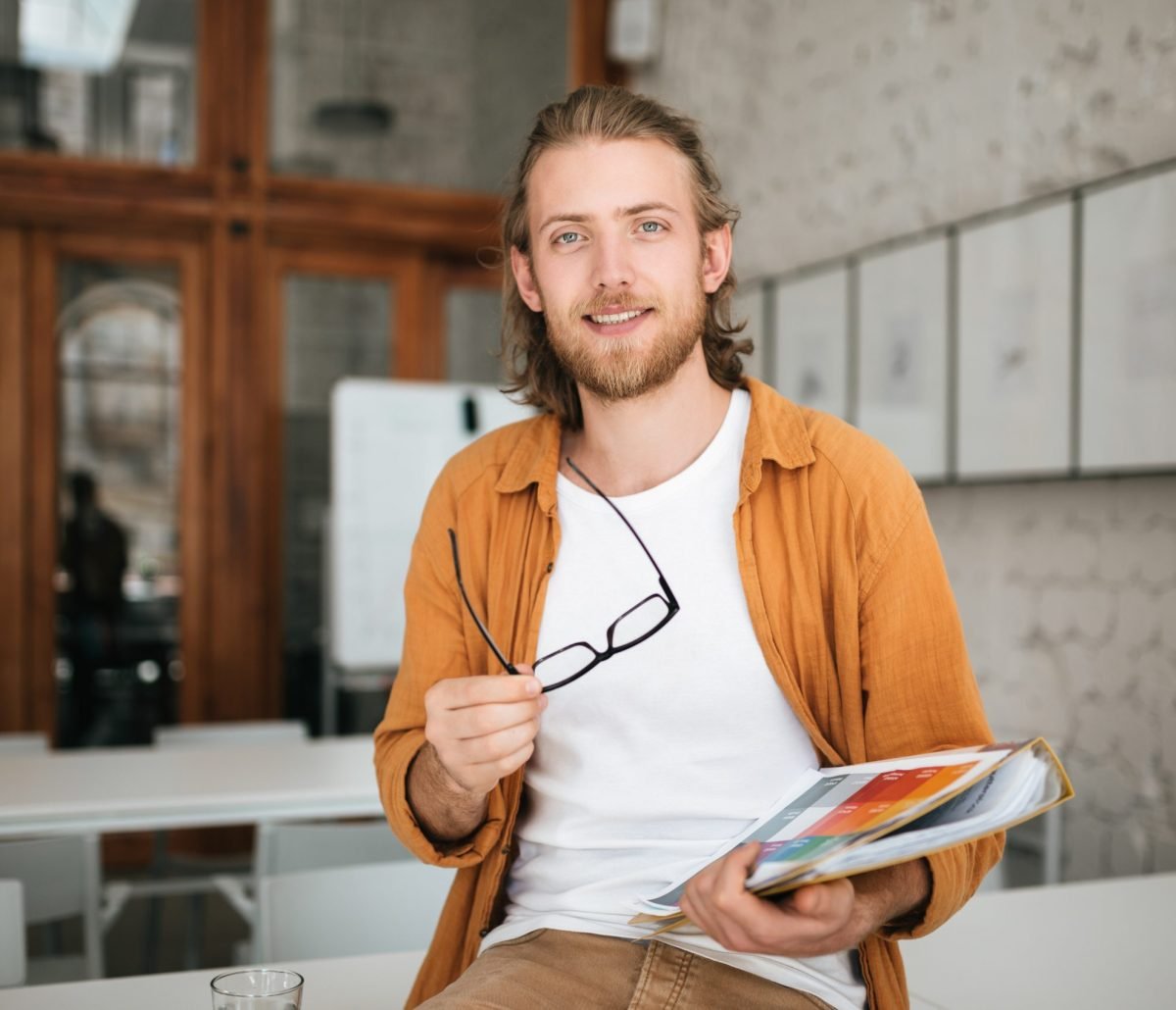 Young man sitting on table in office with glasses and document case in hands. Portrait of smiling boy with blond hair and beard joyfully looking in camera while holding documents in hand