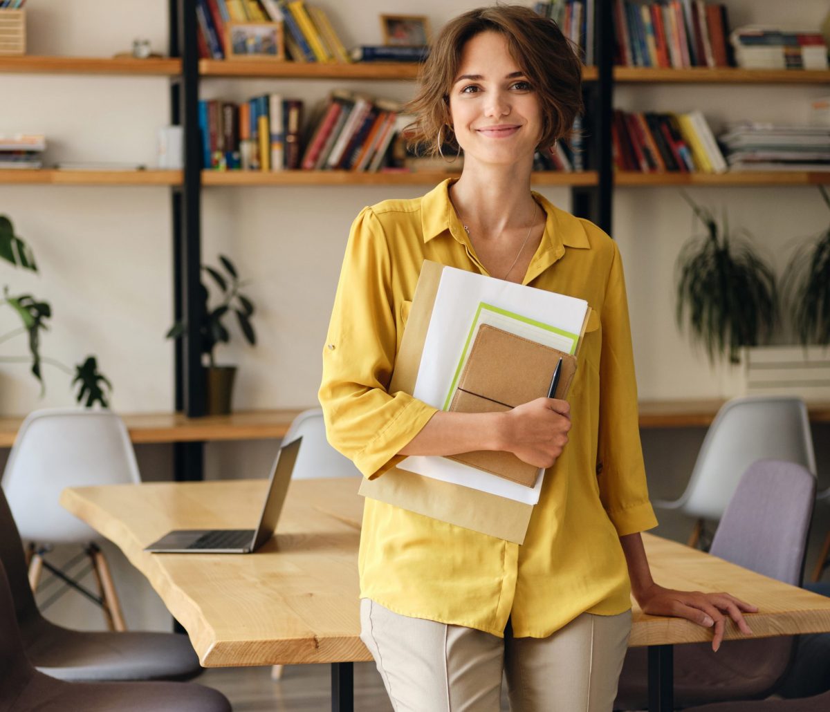 Young smiling woman in yellow shirt leaning on desk with notepad and papers in hand happily looking in camera in modern office