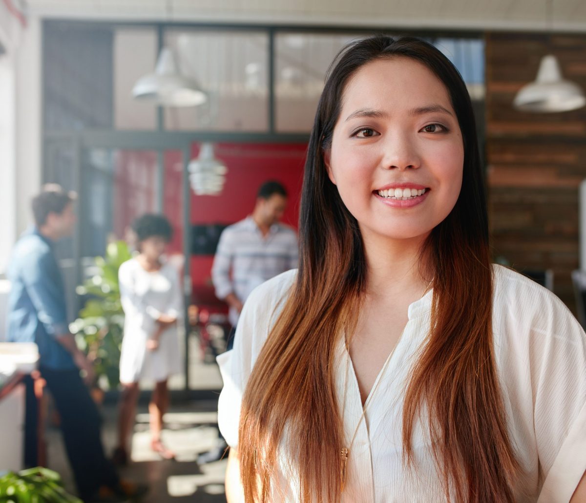Smiling young businesswoman looking at camera and her colleagues are standing in the background. Young asian female creative professional.