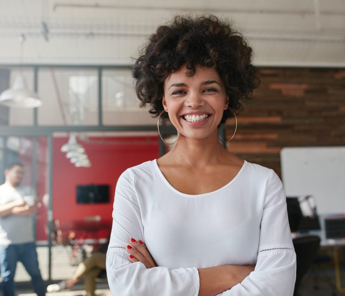 Smiling young woman standing with her arms crossed and looking at camera. She is standing in a modern office with her colleagues in the background.